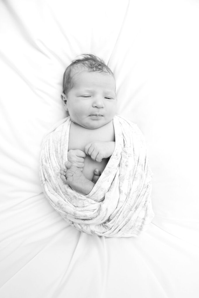 Newborn baby swaddled in a floral wrap lying on white fabric, black and white overhead view