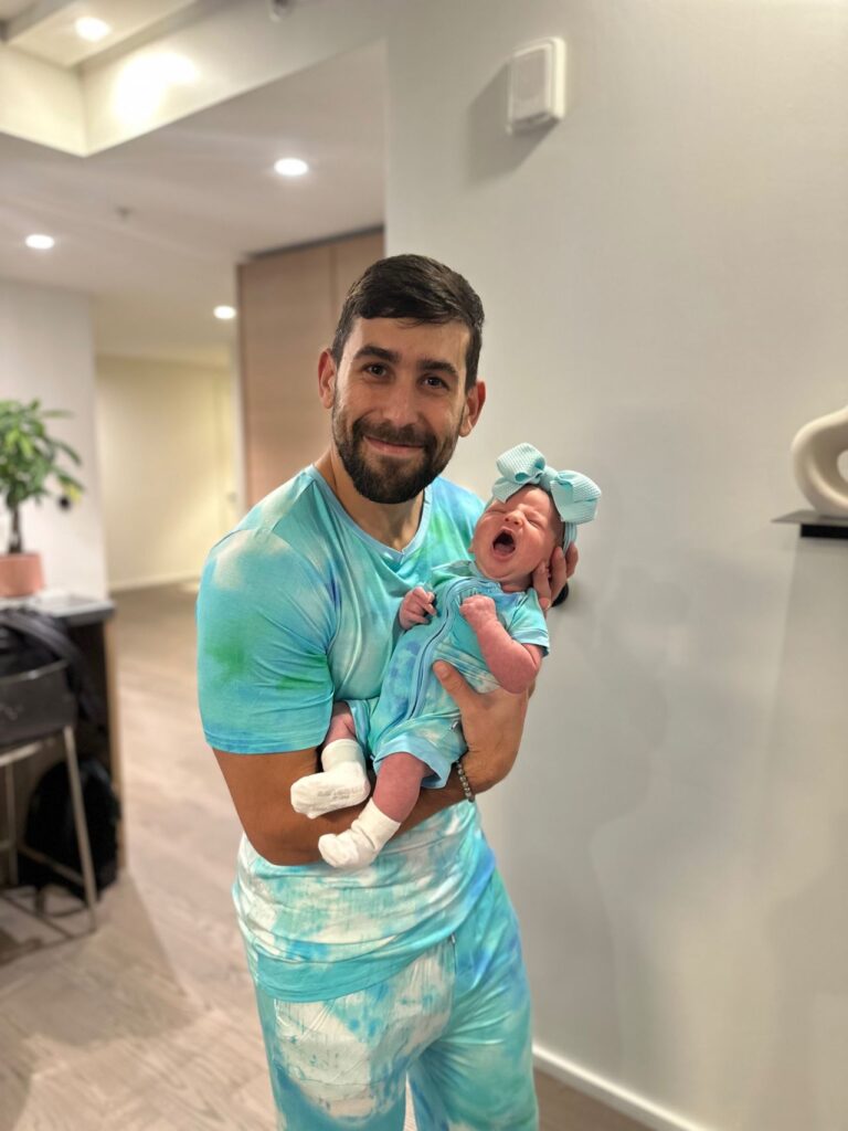 Man holding a newborn girl in matching teal tie-dye outfits, both wearing the same pattern, indoors