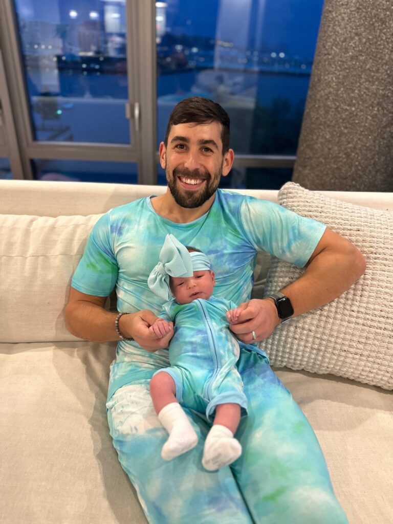 Man and newborn daughter in matching teal tie-dye outfits seated on a white sofa with a city skyline at night in the background